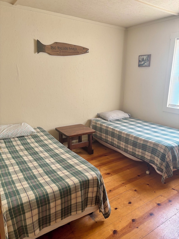 A room with two beds, a small table, a wooden sign on the wall, and a window with blinds.