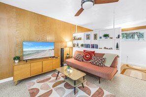 A modern living room with a wooden accent wall, a flat-screen TV, a sofa with patterned cushions, a coffee table, and a bookshelf with various decorative items. - Lido Beach House Family Complex (Sarasota)
