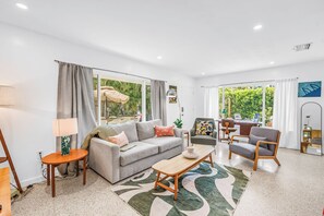 Ein modernes Wohnzimmer mit Sofa, Couchtisch und Sesseln, großen Fenstern mit Vorhängen und Blick ins Grüne.