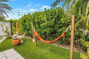 A hammock suspended between wooden posts in a garden with artificial grass, potted plants, and a white building in the background. - Lido Beach House Family Complex (Sarasota)