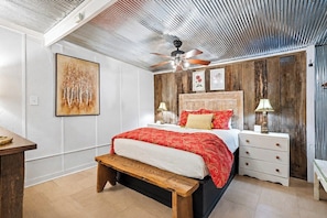 Un dormitorio con cama, mesitas de noche, un banco, ventilador de techo y obras de arte en las paredes.