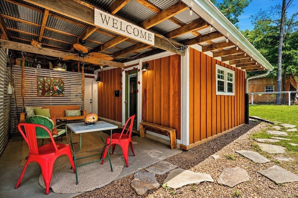 Fachada de madera con un letrero de 'WELCOME', un área de estar al aire libre cubierta con una mesa y sillas, y un camino de piedra que conduce a la entrada.