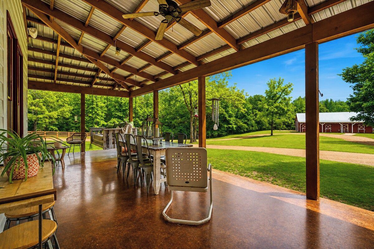 Une terrasse extérieure couverte, dotée de tables et de chaises, d’un ventilateur de plafond et offrant une vue sur un paysage verdoyant avec une grange.