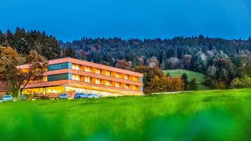 Ein mehrstöckiges Hotel mit flachem Dach, umgeben von einer grünen Landschaft und einem bewaldeten Hügel im Hintergrund.
