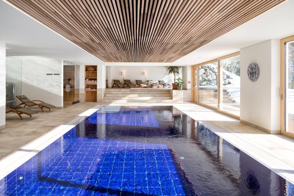Ein moderner Poolbereich im Innenbereich mit blauen Fliesen, Holzdielen an der Decke und einer Lounge-Ecke mit Liegestühlen.