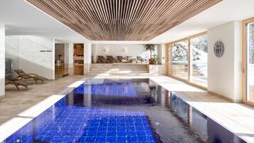 Ein moderner Poolbereich im Innenbereich mit blauen Fliesen, Holzdielen an der Decke und einer Lounge-Ecke mit Liegestühlen.