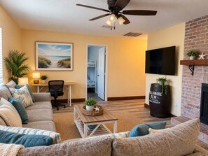 A living room with a sofa, coffee table, and a television. There is a fireplace and a ceiling fan.