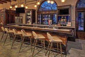 A bar area with a long counter, multiple stools, and a large arched window. - Courtyard by Marriott Kansas City Country Club Plaza (Kansas City)