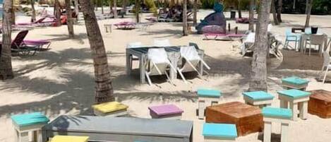 A beach with lounge chairs and tables, some shaded by palm trees.