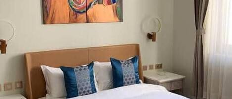 A hotel room with a bed, a nightstand, a painting on the wall, and a window with curtains.
