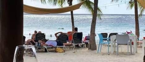 A beachside view with a dining area, palm trees, and beach chairs.
