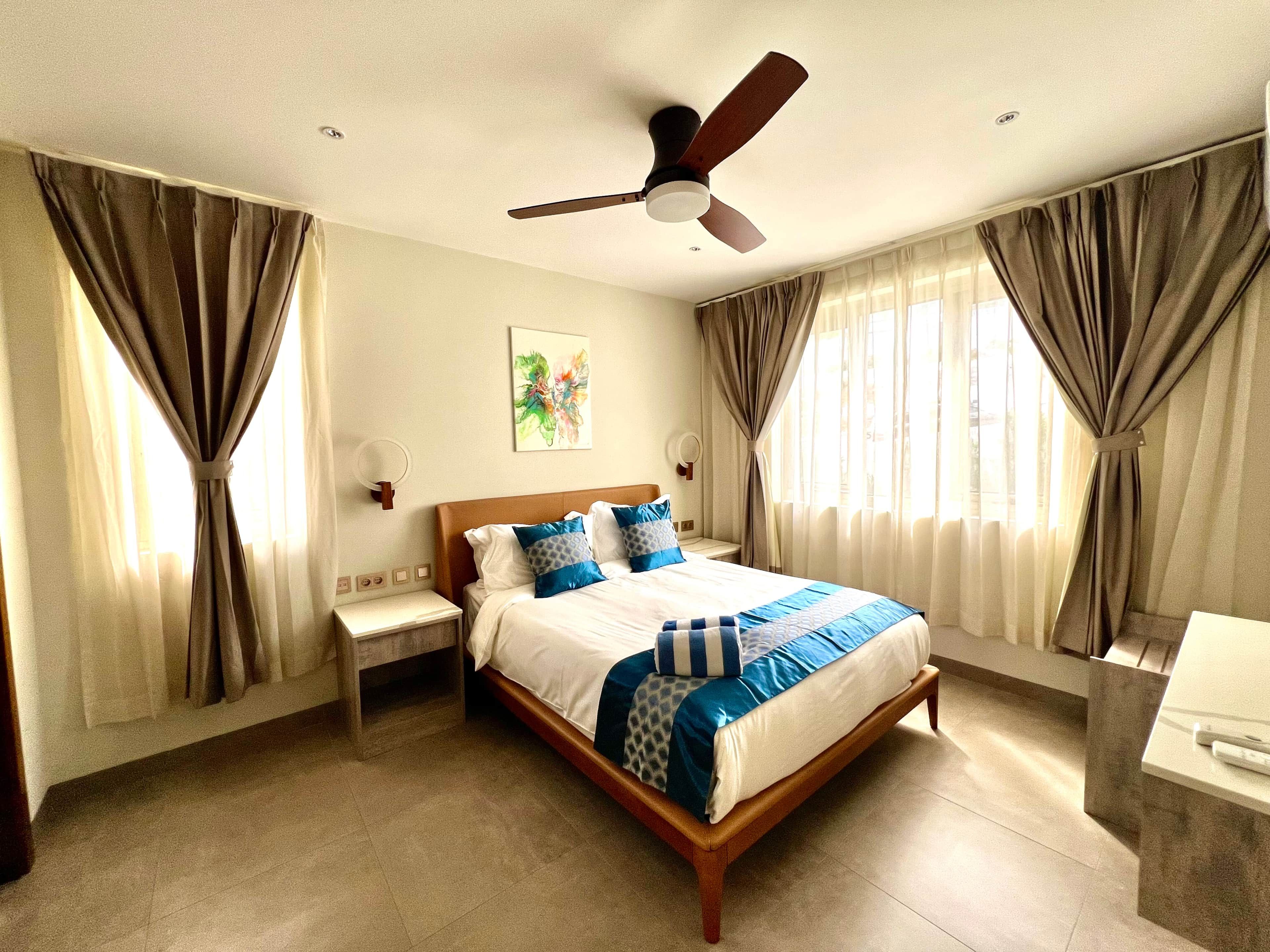 A hotel room with a bed, curtains, a ceiling fan, a bedside table, and a wall-mounted lamp.