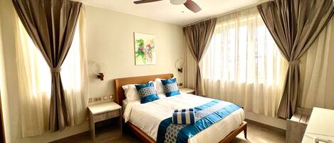 A hotel room with a bed, curtains, a ceiling fan, a bedside table, and a wall-mounted lamp.