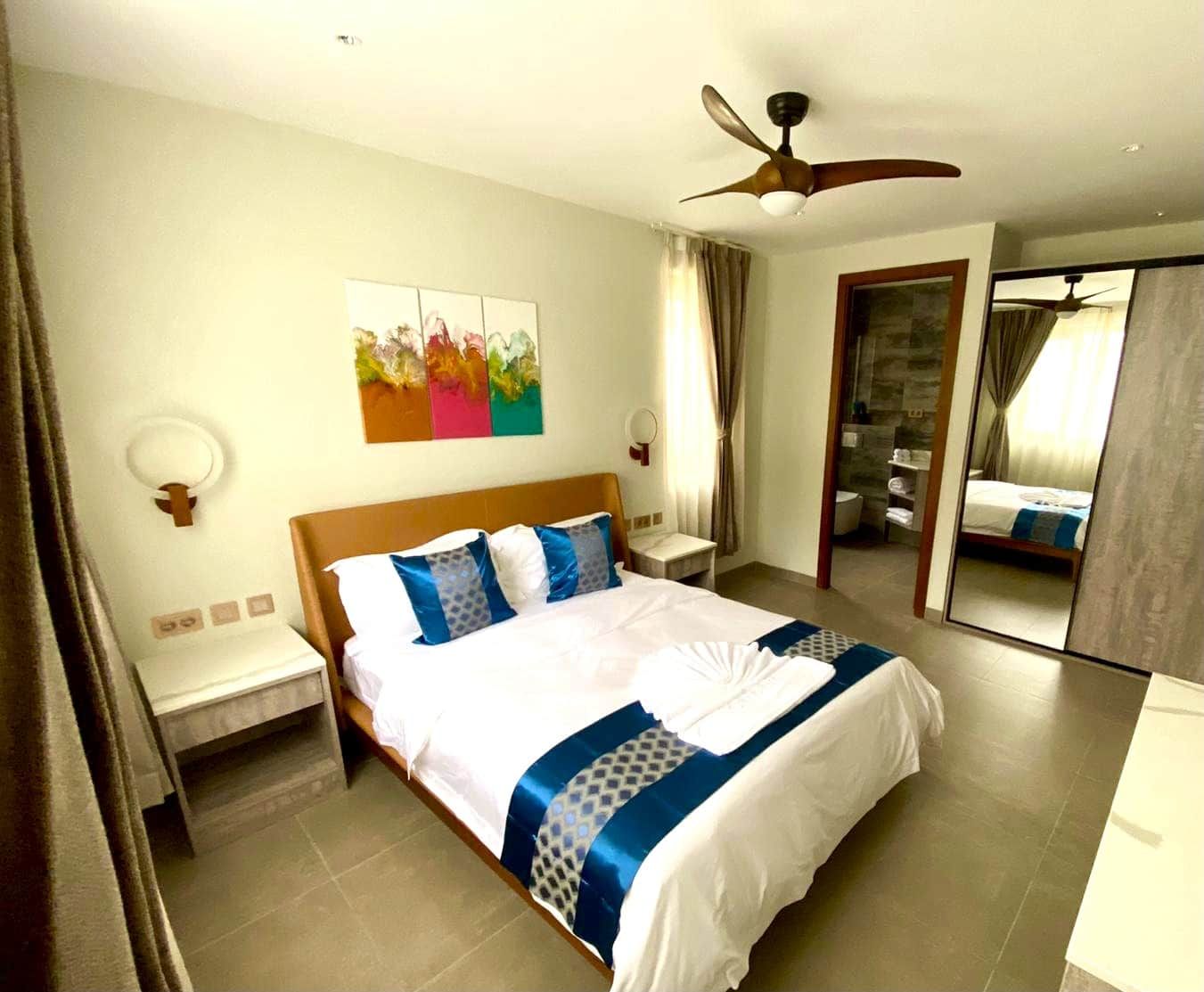Une chambre d’hôtel avec un lit, un ventilateur de plafond, une salle de bain visible à travers une porte ouverte et un tableau accroché au mur.