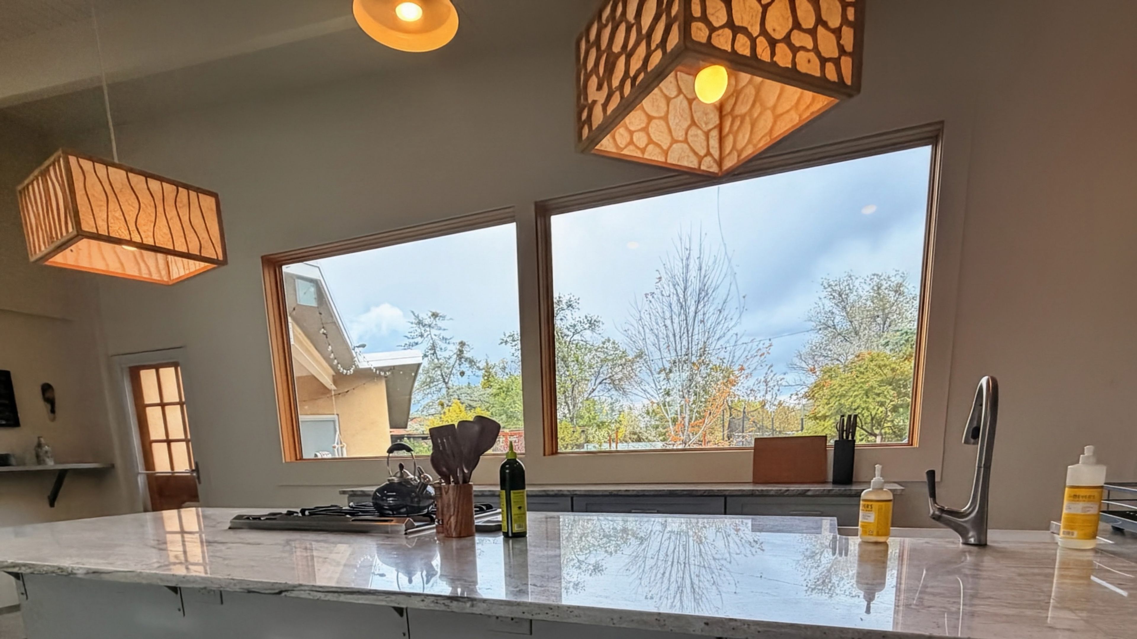 A kitchen with large windows, a marble countertop, and hanging pendant lights.