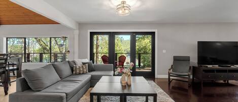 A modern living room with a sectional sofa, a coffee table, and a television. There is a large window providing a view of the outdoors.