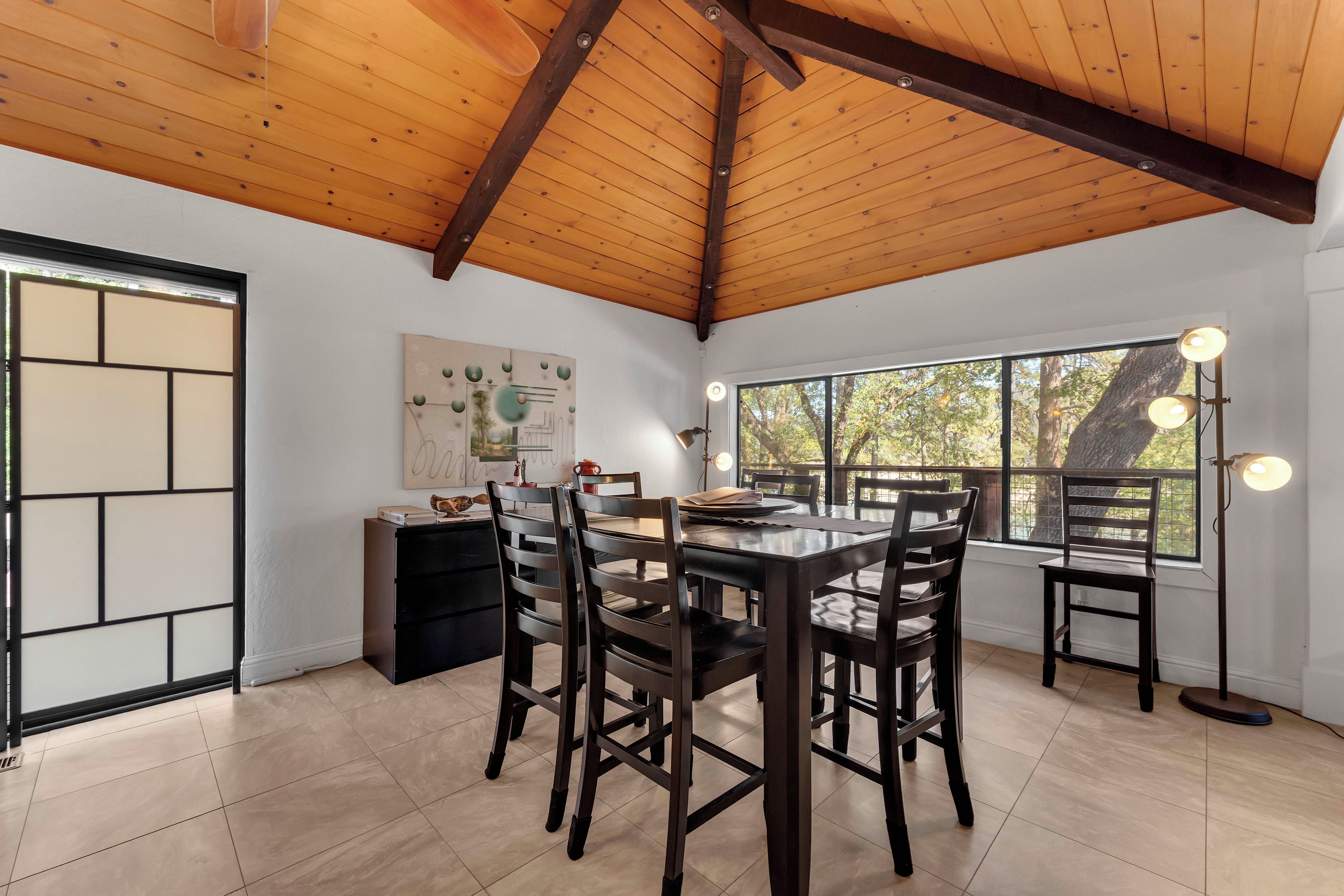A dining area with a wooden ceiling, a table with chairs, a window with lights, and a wall-mounted artwork.