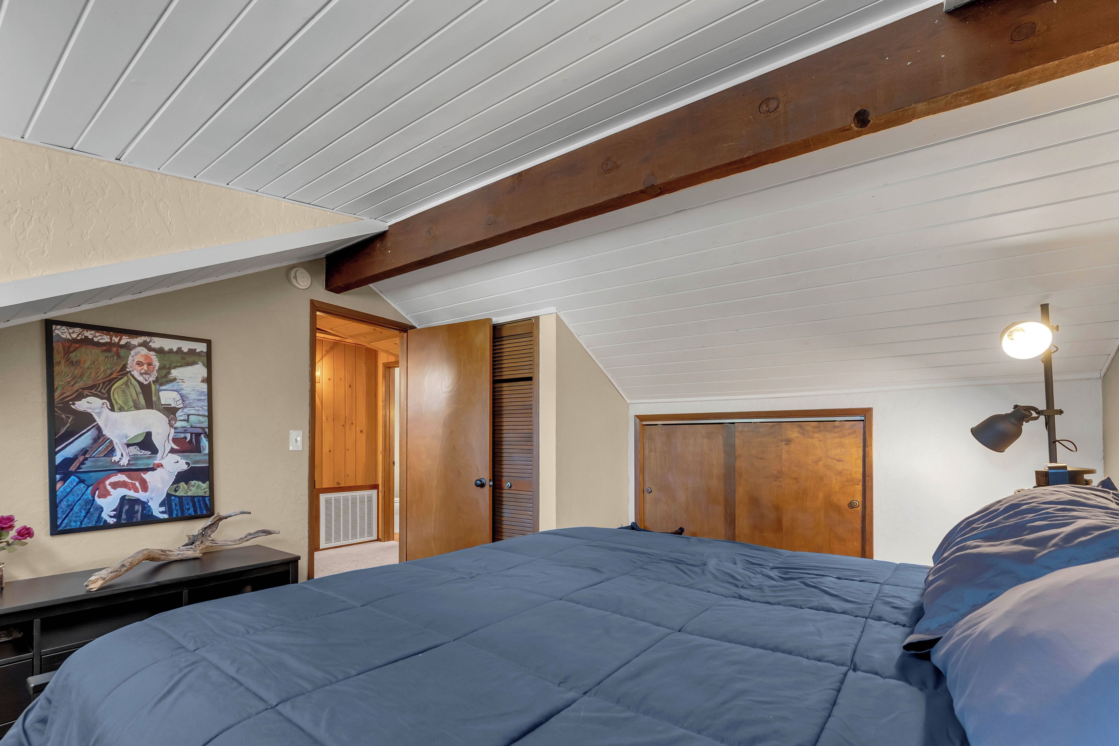 A bedroom with a large bed, a wooden ceiling beam, a door, and a wall-mounted light fixture.