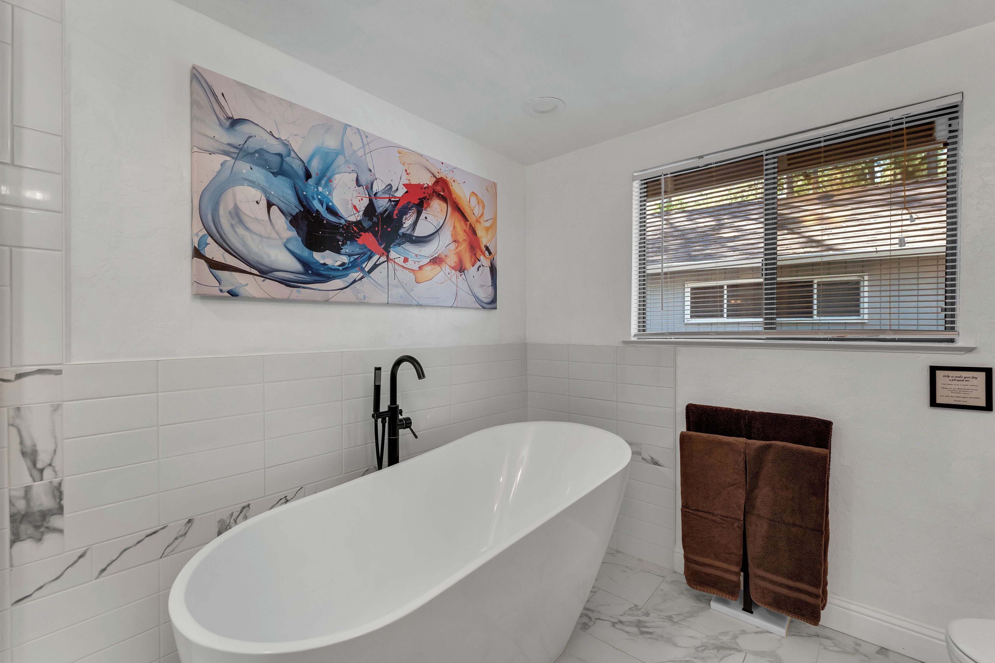A modern bathroom with a freestanding bathtub, a large abstract painting, and a window with blinds.