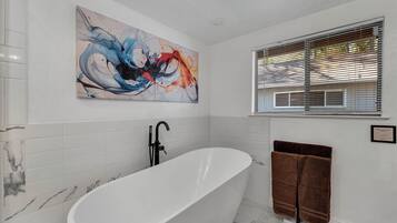A modern bathroom with a freestanding bathtub, a large abstract painting, and a window with blinds.