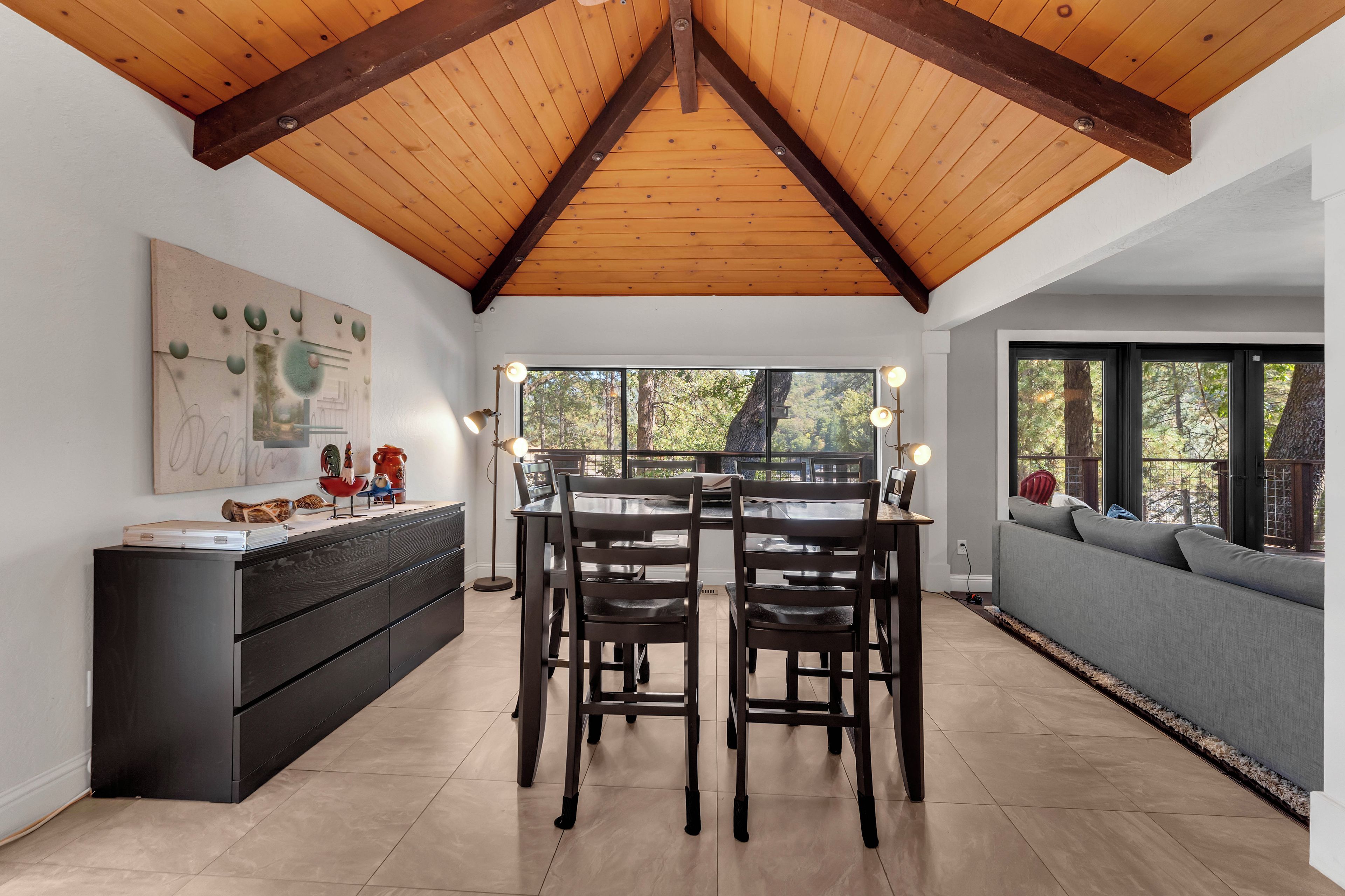 A dining area with a wooden ceiling, a black dining table, and chairs. There is a sofa with a view of trees through large windows.