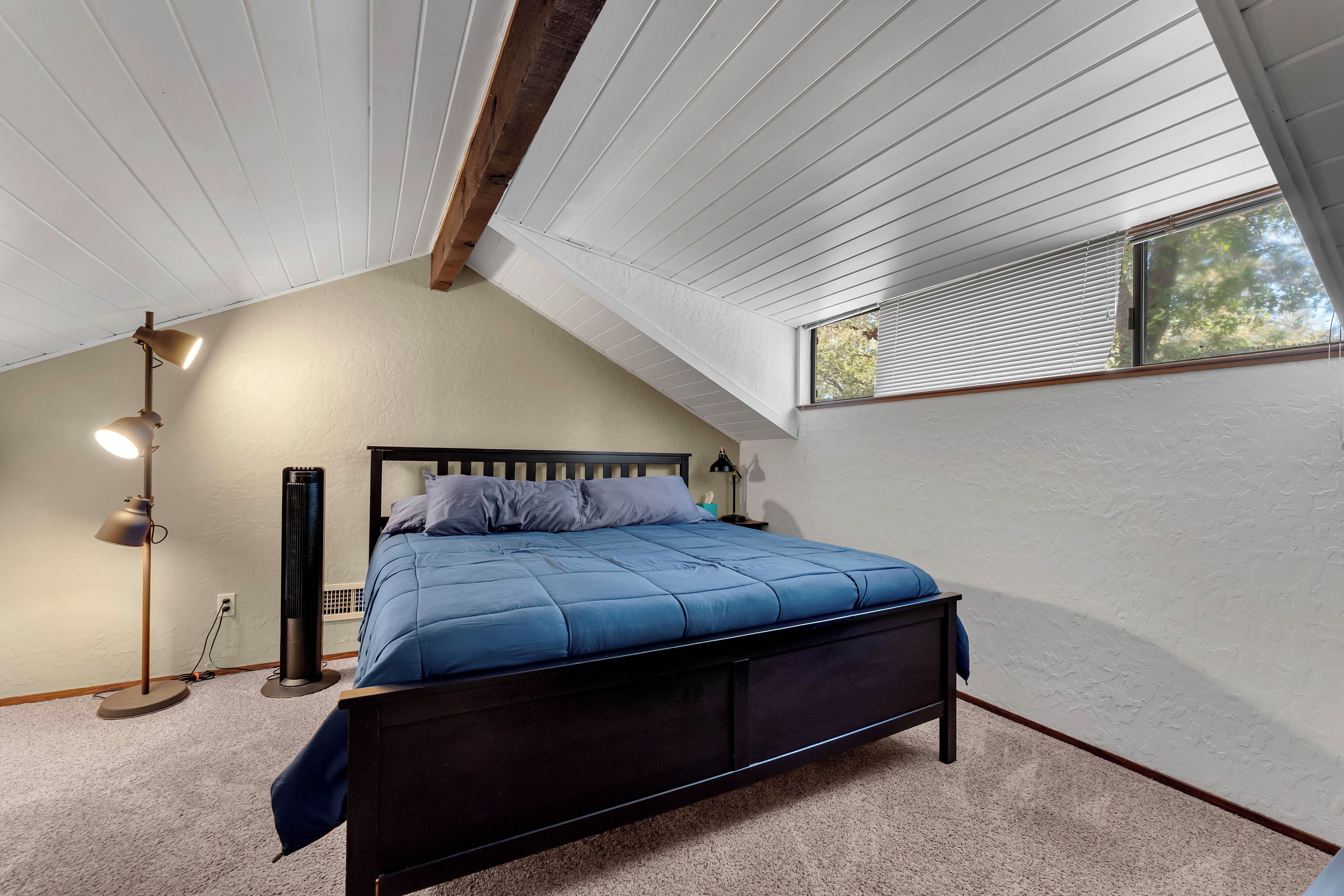 A bedroom with a sloped ceiling, a bed with a blue bedspread, a floor lamp, and a window with blinds.