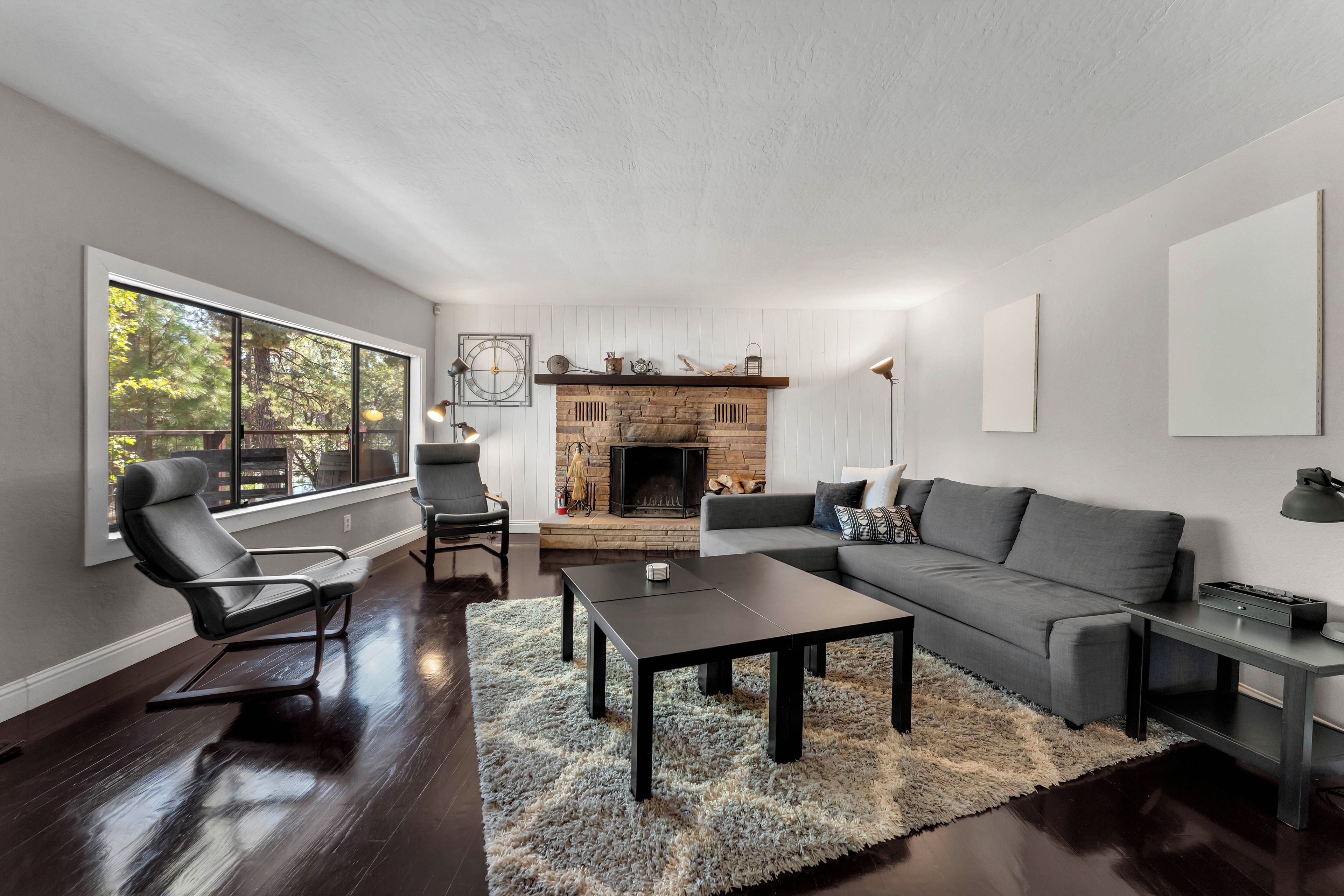 Uma sala de estar moderna com lareira, sofá, mesa de centro e uma grande janela com vista para árvores.