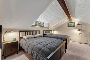 Ein Schlafzimmer mit schräg stehender Decke, einem Bett mit grauer Tagesdecke, Nachttischlampen, einem Nachttisch und einem gerahmten Bild an der Wand.