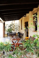 Un patio exterior con una mesa y sillas de madera, rodeado de plantas y un muro de piedra.