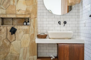 A bathroom with a stone wall, a white tiled backsplash, a round mirror, a white sink, and a wooden cabinet. - Villa SolaFide - Stunning 2BR Pool Villa (Ungasan)