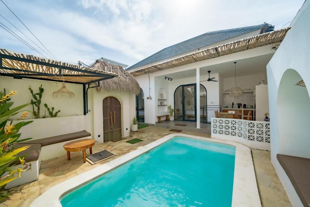 Zona de piscina con techo de paja, banco de madera y pared blanca con una puerta.
