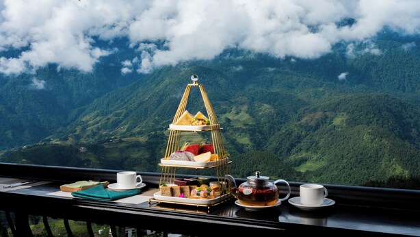 A table with a tiered tray of food, two cups, and a teapot, set against a mountainous backdrop. - Holiday Sapa (Sa Pa)
