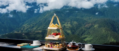 A table with a tiered tray of food, two cups, and a teapot, set against a mountainous backdrop.