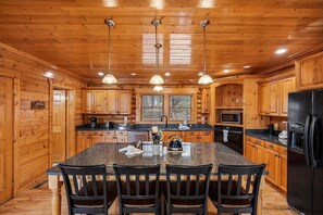 Ein Interieur einer Holzhütte mit einer Küche, die sich durch Granit Arbeitsplatten, eine zentrale Kochinsel mit Stühlen und einen schwarzen Kühlschrank auszeichnet.