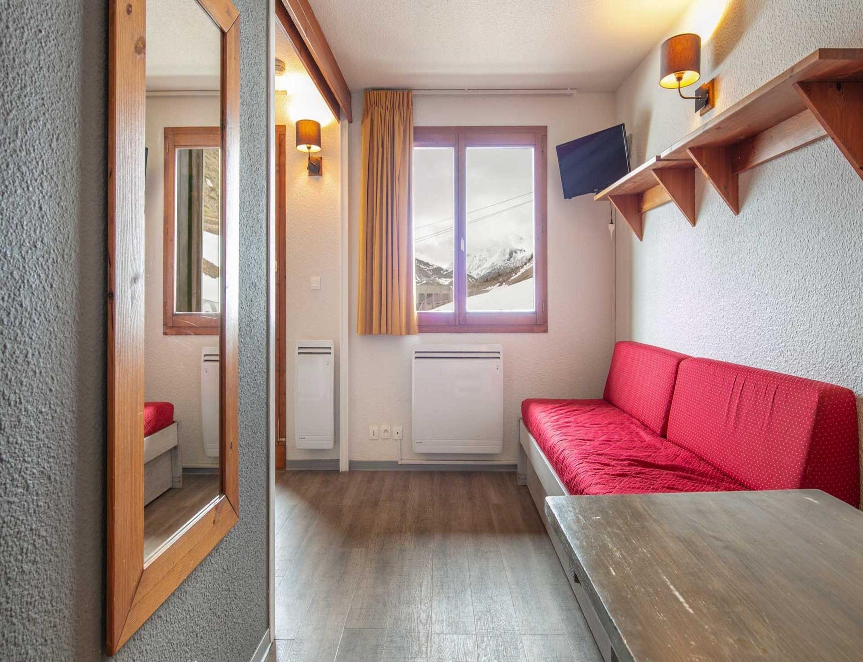 A compact room with a red sofa, a wooden table, a mirror, a window with curtains, and a mounted TV.