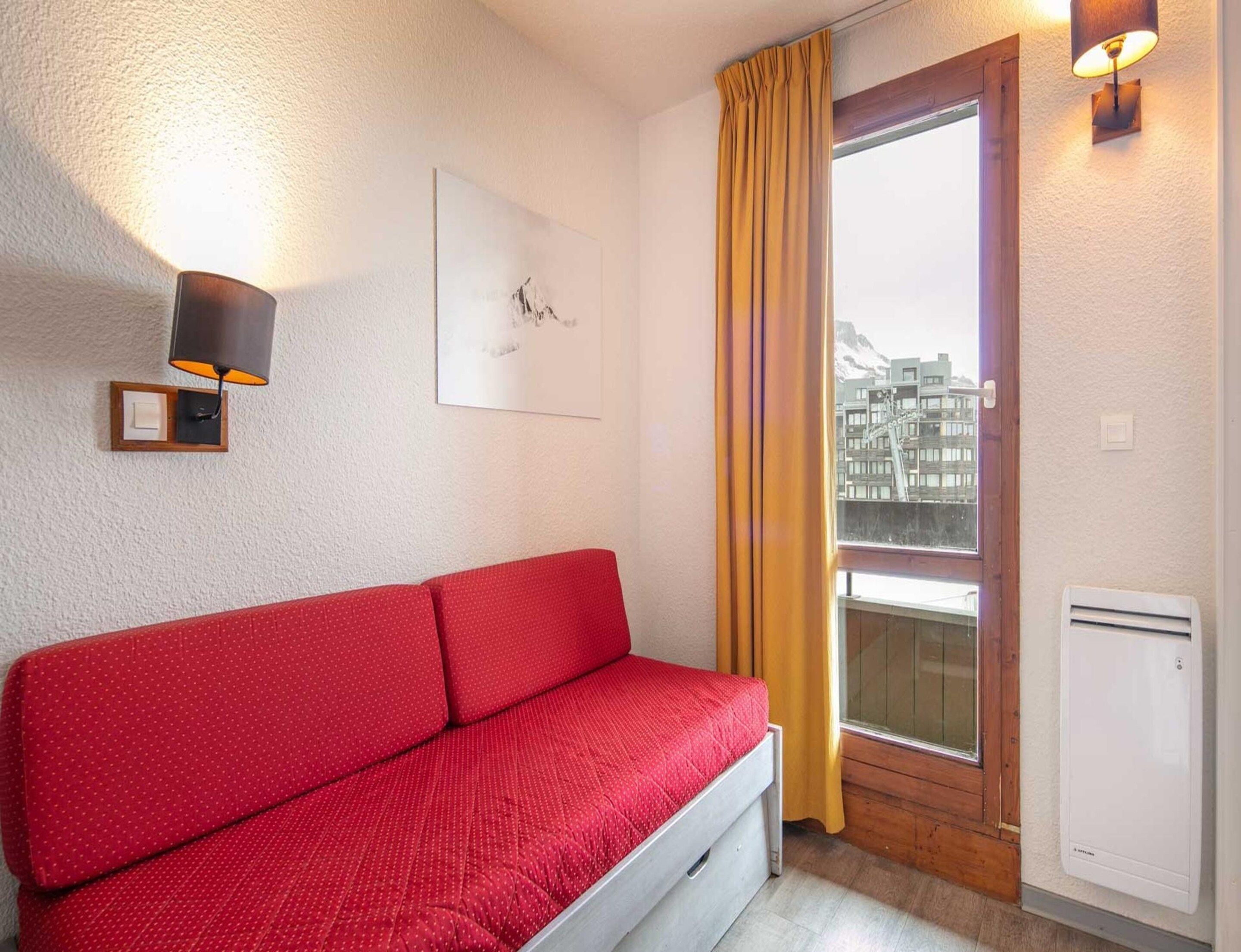 A red sofa in a room with a window, a wall-mounted light, and a framed picture.