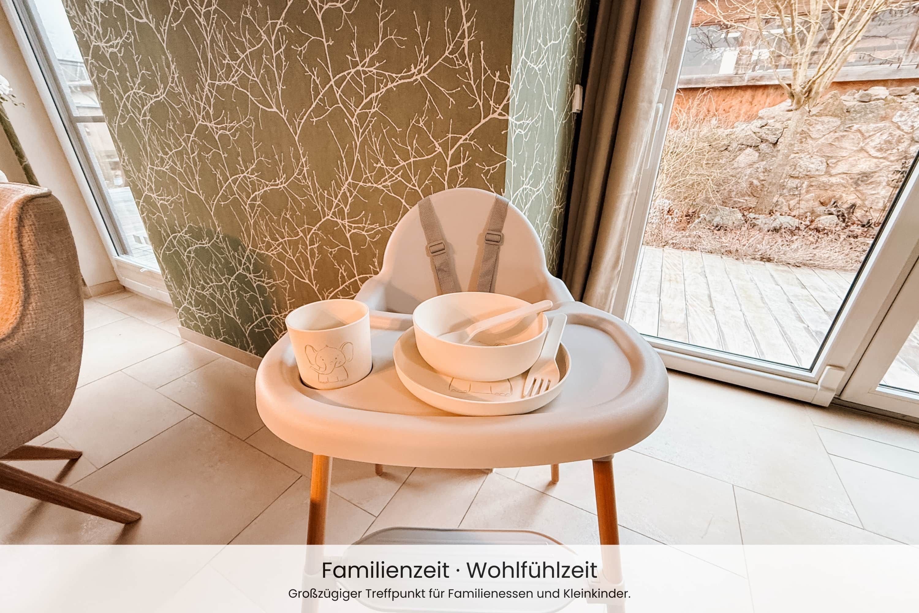 A high chair with a tray, a cup, and a bowl, positioned near a window with a view of a garden.
