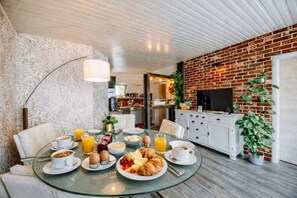 Una escena de desayuno con una mesa de vidrio, sillas blancas y una pared de ladrillo.
