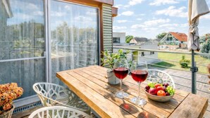 Een houten tafel met twee wijnglazen en een schaal fruit op een balkon, met uitzicht op huizen en bomen.