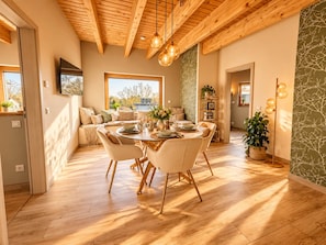 A dining area with a wooden ceiling, a round table with chairs, and a view of the outdoors through large windows.