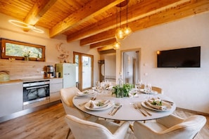 A dining area with a round table set for four, wooden ceiling beams, a television mounted on the wall, and a kitchen with modern appliances.