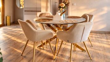 A dining area with a round glass table, four upholstered chairs with gold legs, and a vase with flowers on the table.