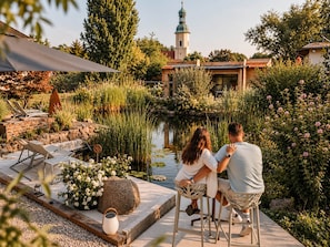 A couple sitting at a table by a pond with a gazebo and lounge chairs in the background.