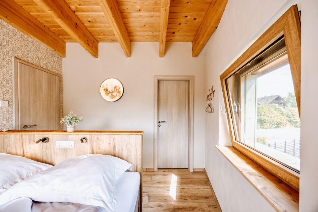 A bedroom with a wooden ceiling, a large window, a bed with white bedding, a bedside table, and a wall-mounted light fixture.