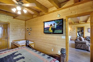 Ein Holzhütten-Innenraum mit einem an der Wand montierten Fernseher, einer Badewanne und einem Bett mit einem gemusterten Bettbezug.
