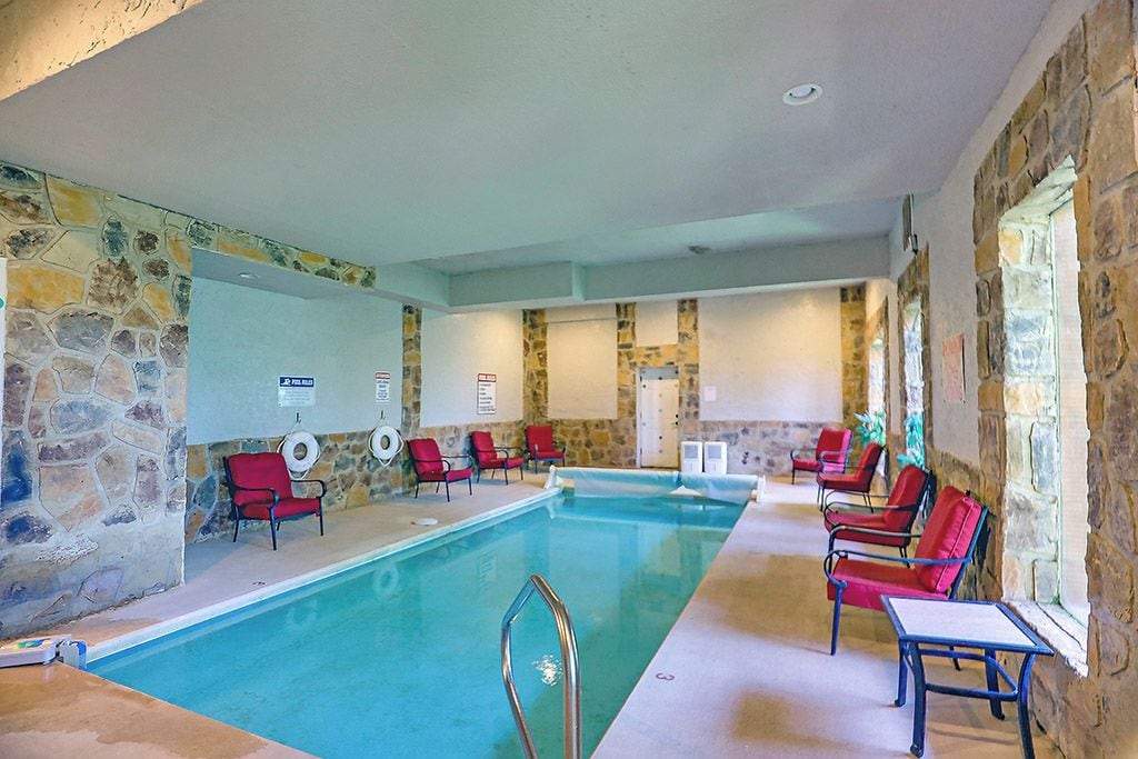 An indoor swimming pool with stone walls, red chairs, and a poolside table.