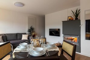 Ein modernes Wohnzimmer mit Kamin, einer Couch, einem Esstisch für vier Personen und einem an der Wand befestigten Fernseher.