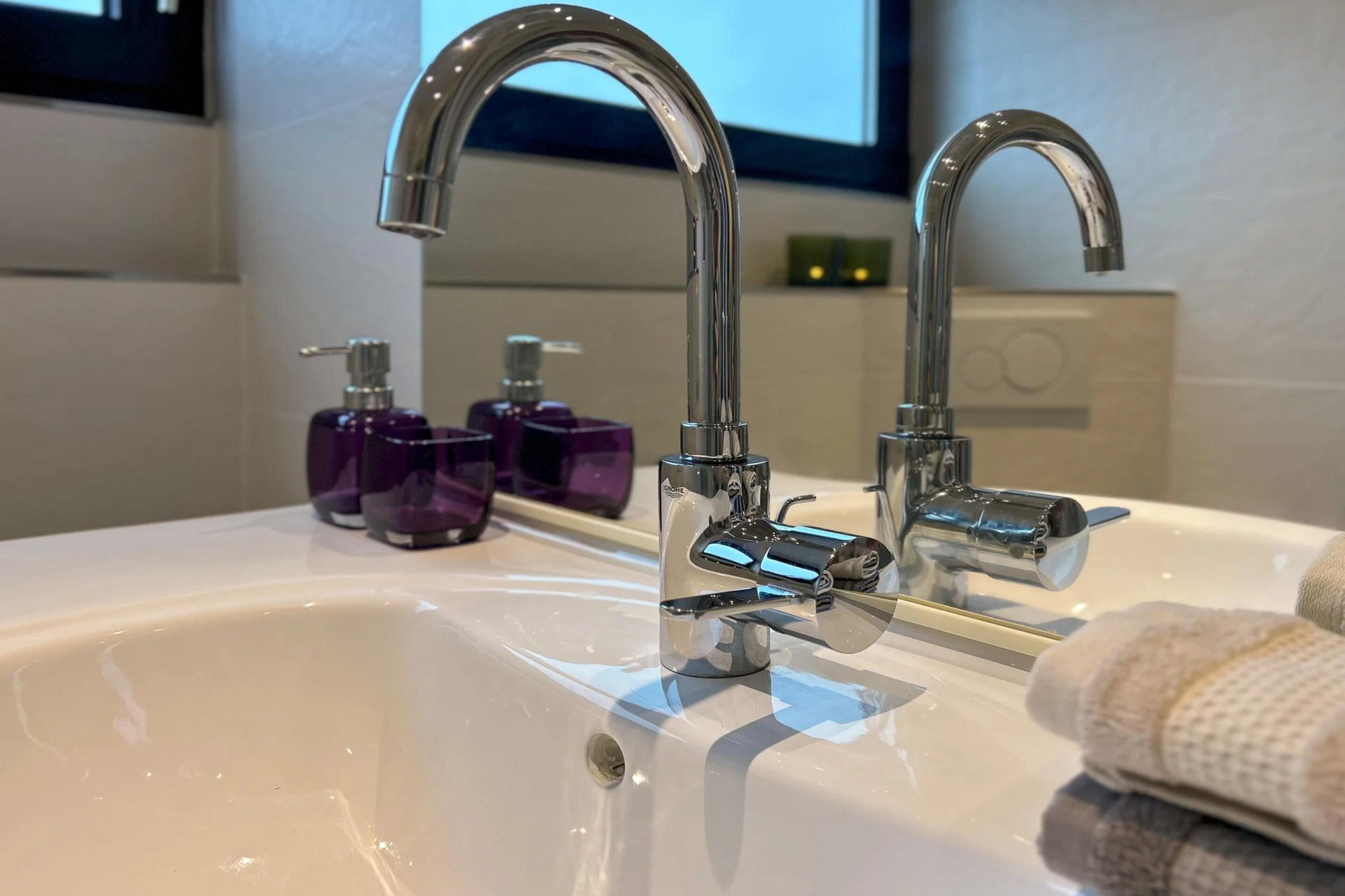 Une salle de bain avec un lavabo blanc, deux robinets chromés et des distributeurs de savon violets.