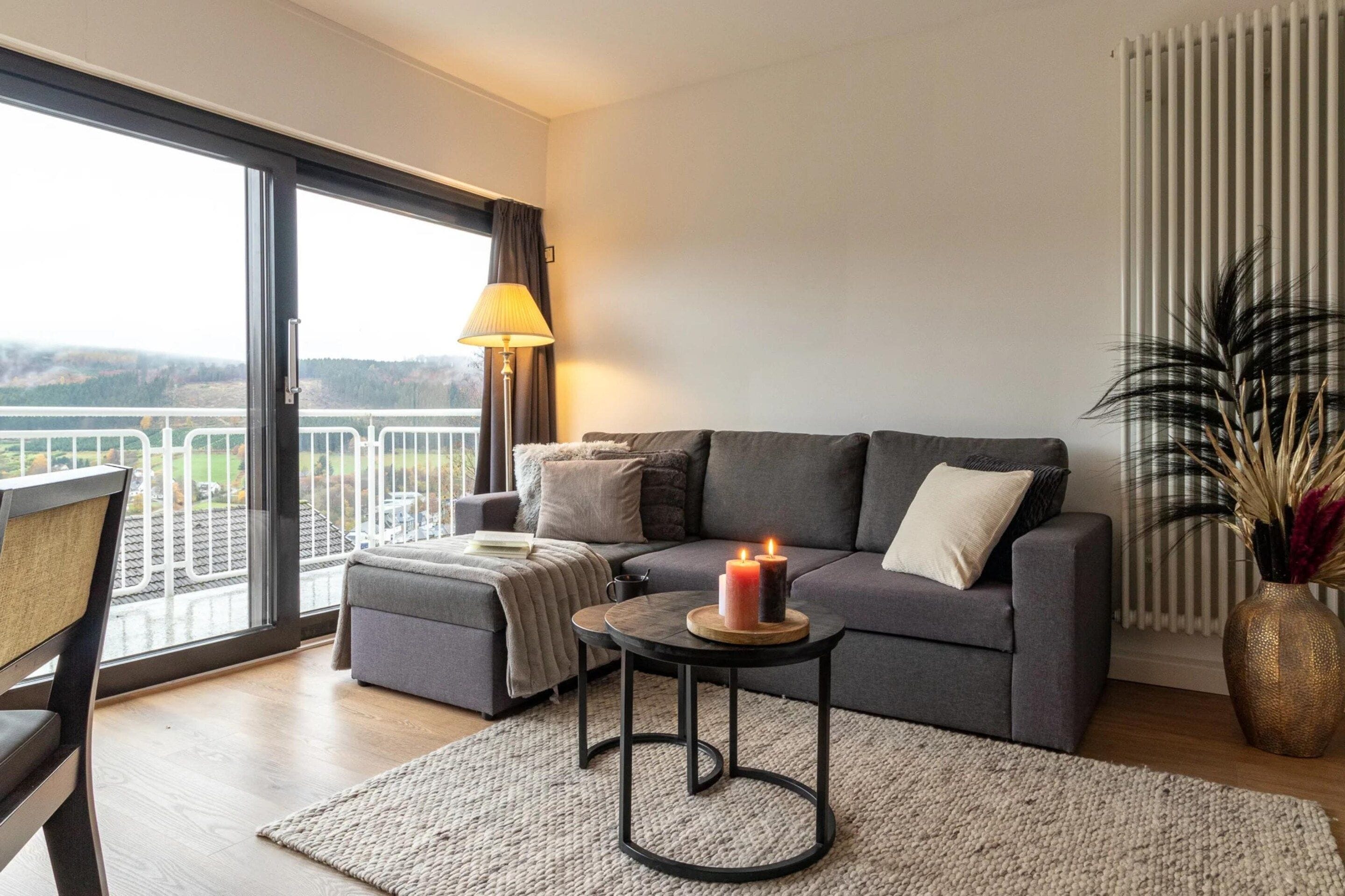 Een moderne woonkamer met een grijze bank, een klein rond tafeltje met kaarsen, uitzicht door een groot raam, en een bloempot met plant.