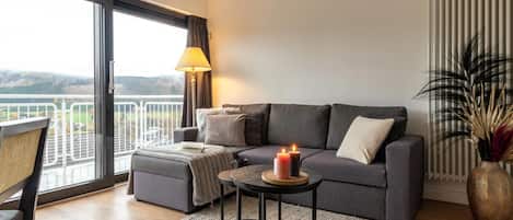 Een moderne woonkamer met een grijze bank, een klein rond tafeltje met kaarsen, uitzicht door een groot raam, en een bloempot met plant.
